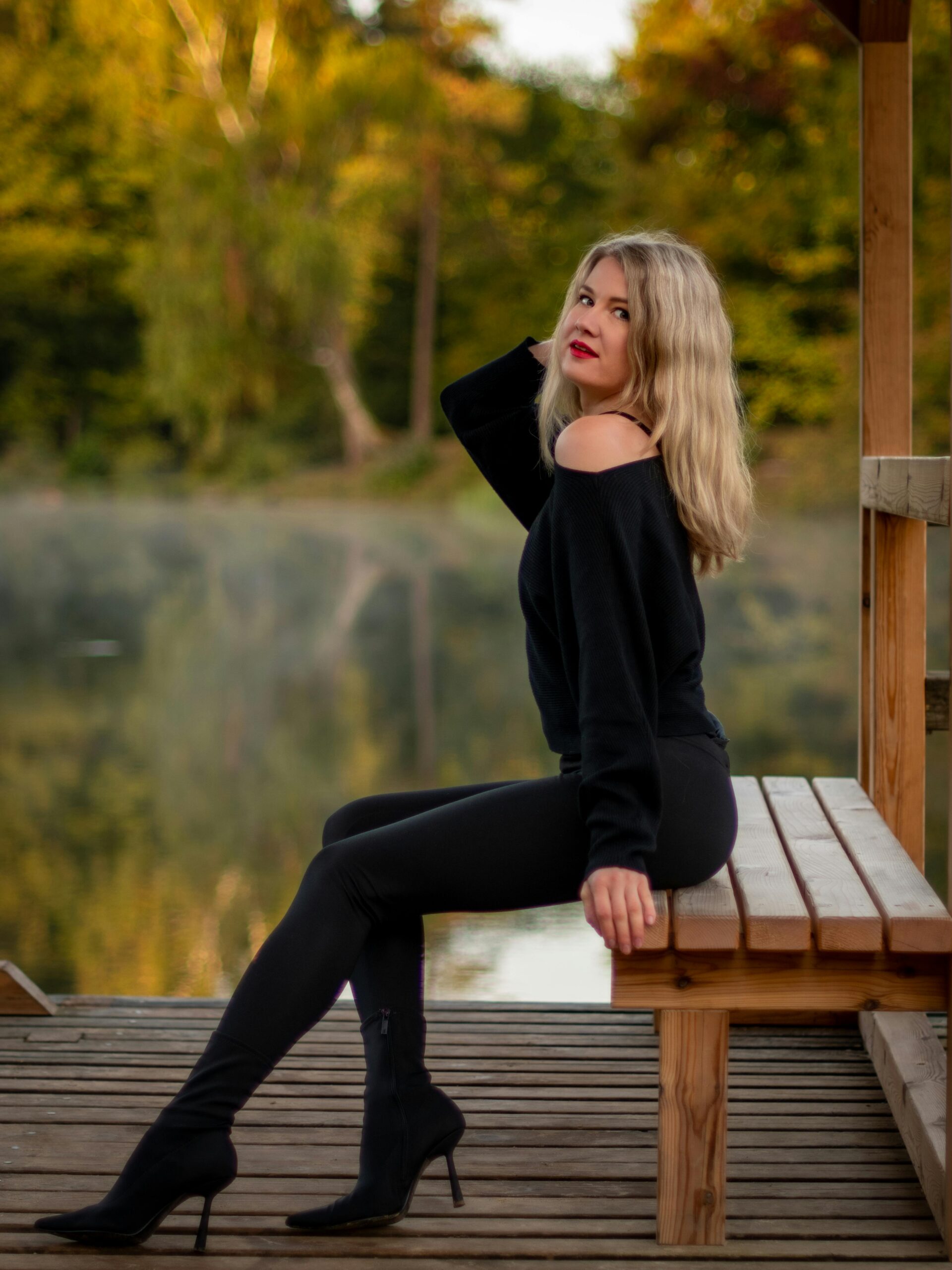 Elegant woman in black outfit posing on a bench by the lake during a serene autumn day in Vilnius, Lithuania.