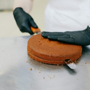 A baker in gloves skillfully slices a chocolate cake with a spatula on a metal table.